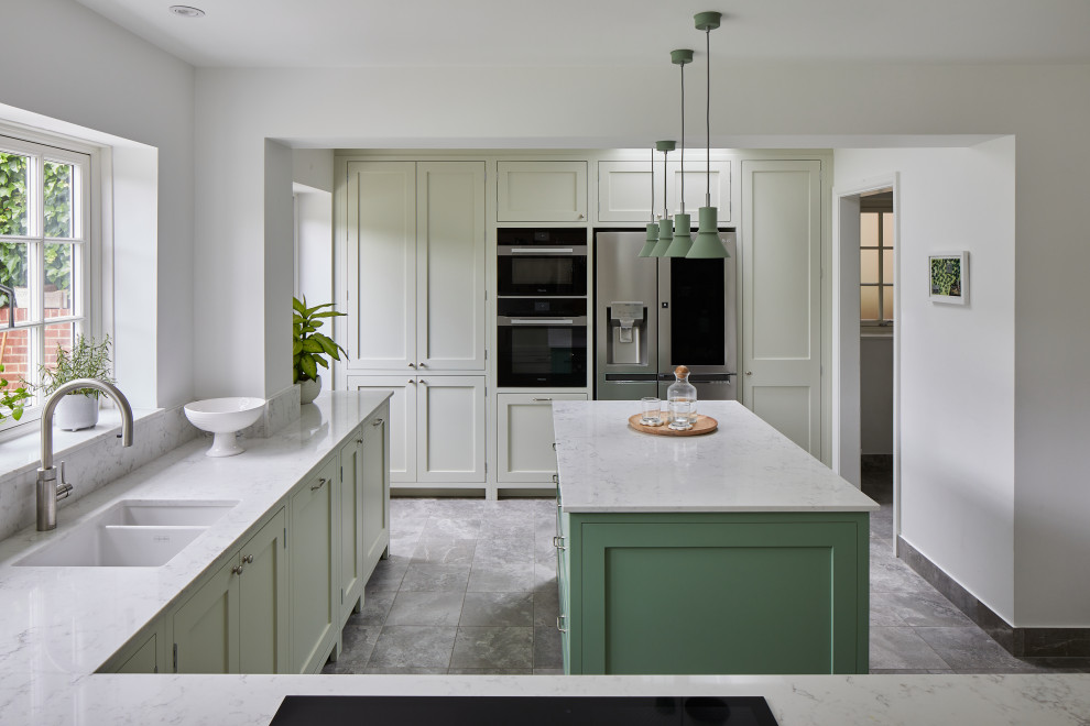 Mid-sized transitional l-shaped separate kitchen in London with a drop-in sink, shaker cabinets, green cabinets, quartzite benchtops, grey splashback, engineered quartz splashback, stainless steel appliances, ceramic floors, with island, grey floor and grey benchtop.