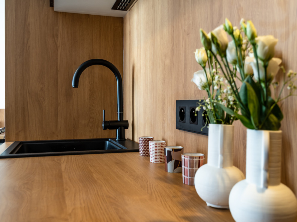 Cette image montre une petite cuisine ouverte linéaire et noire et bois design en bois clair avec un évier posé, un placard à porte plane, un plan de travail en bois, une crédence en bois, un électroménager noir, moquette et un sol gris.