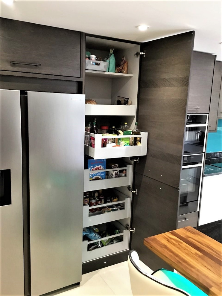 Photo of a large modern l-shaped open plan kitchen in Other with a submerged sink, flat-panel cabinets, grey cabinets, granite worktops, blue splashback, glass sheet splashback, black appliances, porcelain flooring, an island, beige floors, black worktops and a feature wall.