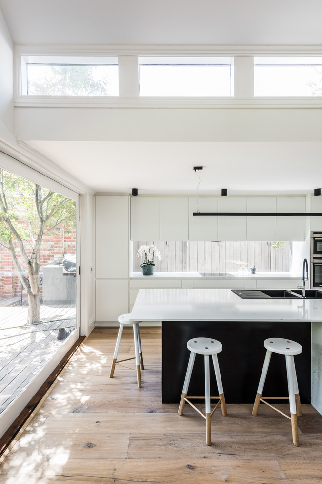 Inspiration for a modern galley kitchen in Melbourne with a drop-in sink, flat-panel cabinets, white cabinets, window splashback, stainless steel appliances, medium hardwood floors, with island, brown floor and white benchtop.