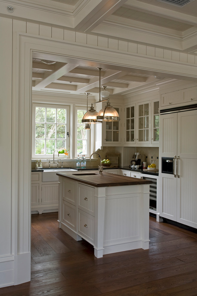 Inspiration for a beach style u-shaped kitchen in Other with a farmhouse sink, recessed-panel cabinets, white cabinets, wood benchtops, beige splashback, panelled appliances, medium hardwood floors, with island and brown floor.