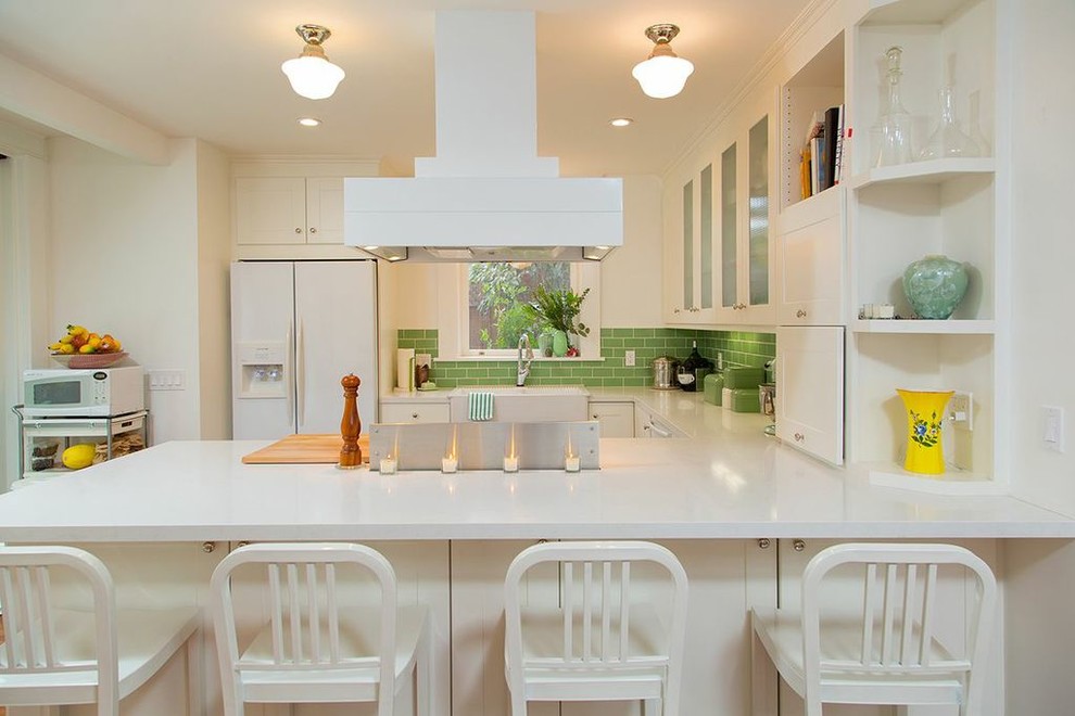 This is an example of a transitional u-shaped kitchen in San Diego with a farmhouse sink, glass-front cabinets, white cabinets, quartz benchtops, green splashback, subway tile splashback and white appliances.