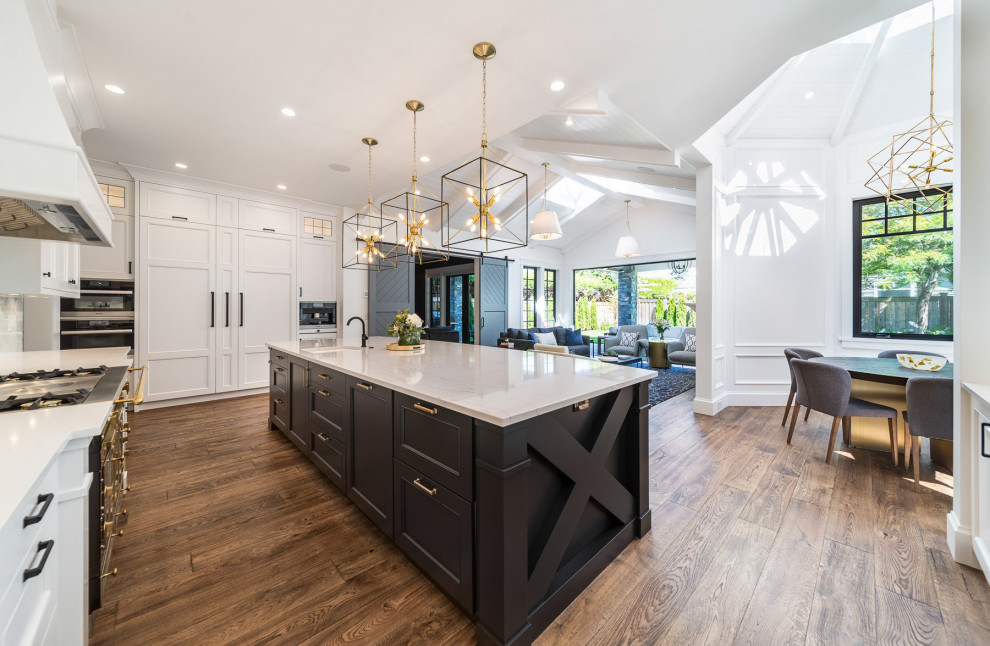 Photo of a large arts and crafts open plan kitchen in Vancouver with a farmhouse sink, shaker cabinets, white cabinets, quartz benchtops, white splashback, porcelain splashback, black appliances, dark hardwood floors, with island, brown floor, white benchtop and timber.