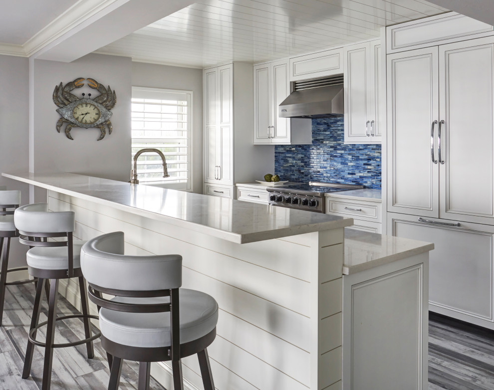 Inspiration for a mid-sized beach style galley open plan kitchen with an undermount sink, recessed-panel cabinets, white cabinets, quartz benchtops, blue splashback, glass tile splashback, panelled appliances, porcelain floors, a peninsula, grey floor, white benchtop and timber.