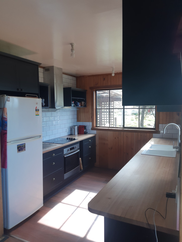Design ideas for a small country galley separate kitchen in Hobart with a farmhouse sink, recessed-panel cabinets, black cabinets, wood benchtops, white splashback, subway tile splashback, stainless steel appliances and vinyl floors.