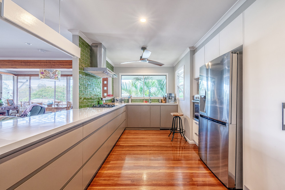 Large contemporary u-shaped kitchen in Brisbane with flat-panel cabinets, beige cabinets, green splashback, ceramic splashback, stainless steel appliances, medium hardwood floors, a peninsula, brown floor and white benchtop.