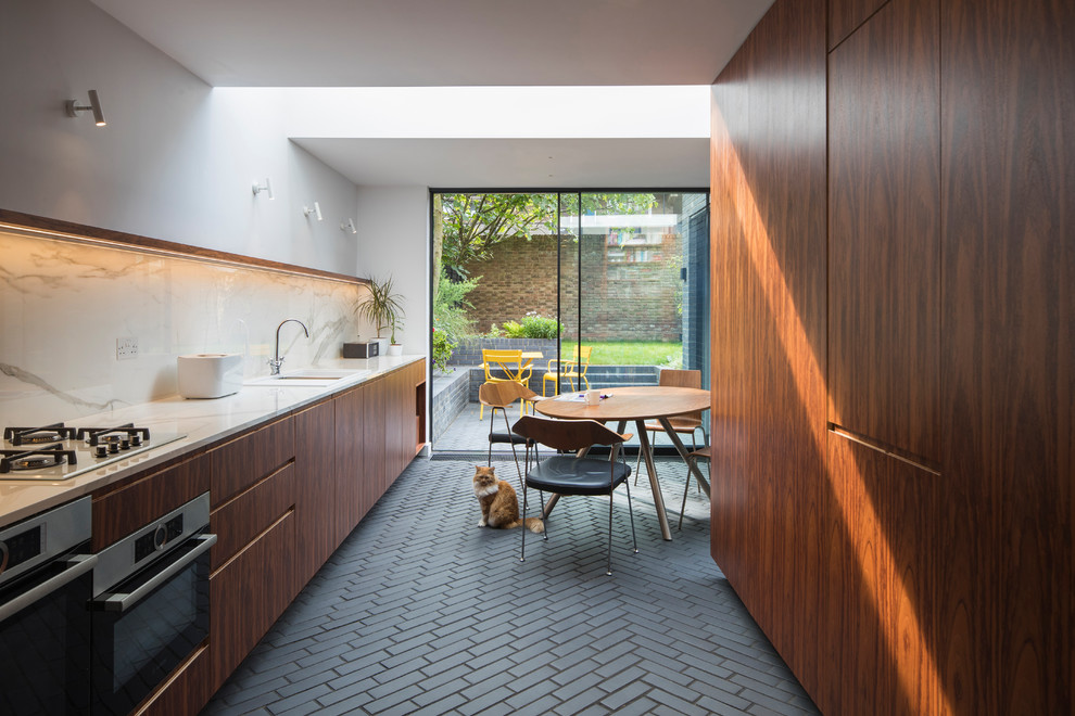 Inspiration for a contemporary single-wall eat-in kitchen in London with flat-panel cabinets, medium wood cabinets, marble benchtops, white splashback, marble splashback, brick floors, no island, black floor, an undermount sink and black appliances.