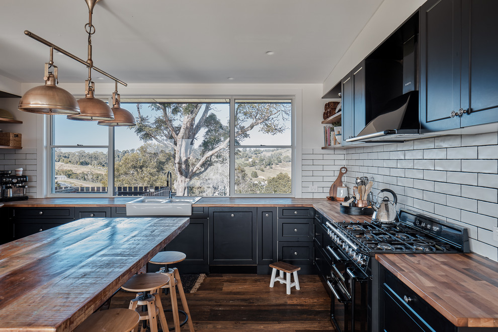 Design ideas for a country l-shaped kitchen in Sunshine Coast with a farmhouse sink, recessed-panel cabinets, black cabinets, wood benchtops, white splashback, subway tile splashback, black appliances, dark hardwood floors, with island, brown floor and brown benchtop.