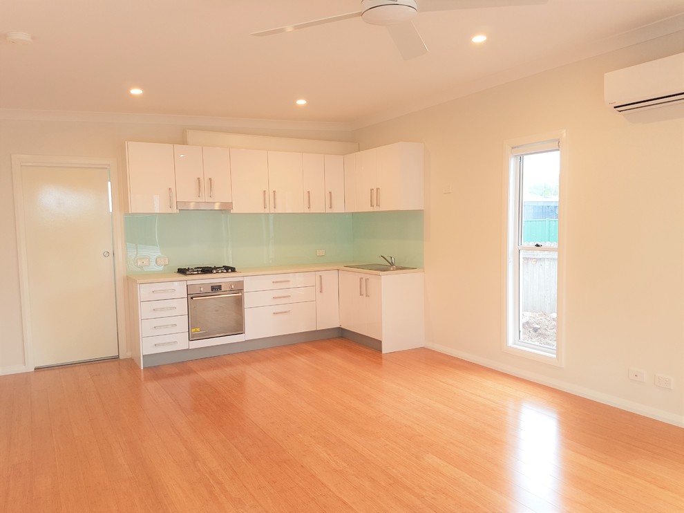 This is an example of a small modern l-shaped open plan kitchen in Sydney with a single-bowl sink, flat-panel cabinets, white cabinets, laminate benchtops, green splashback, glass sheet splashback, stainless steel appliances, bamboo floors and no island.