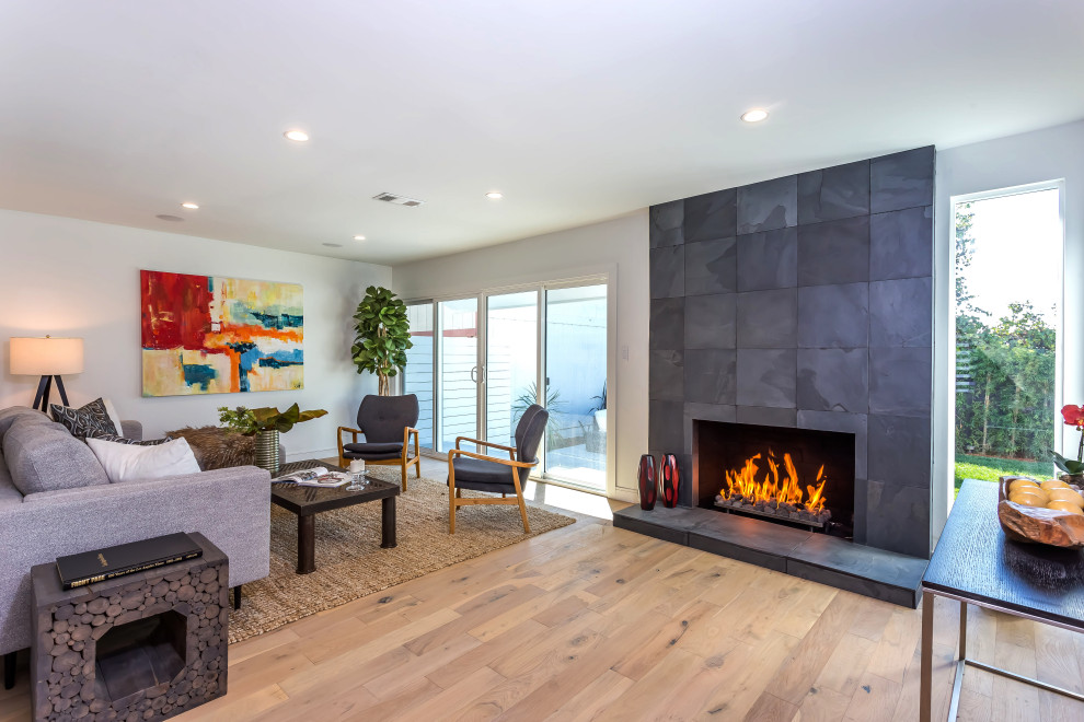 This is an example of a small country open concept living room in Los Angeles with white walls, light hardwood floors, a standard fireplace, a tile fireplace surround, a wall-mounted tv and beige floor.