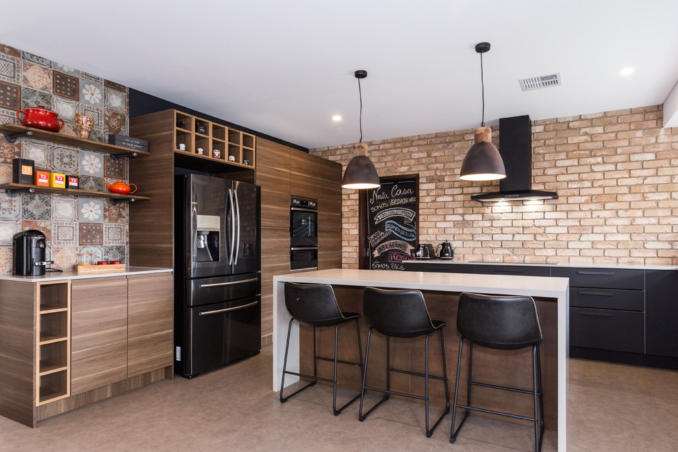 Inspiration for a contemporary l-shaped kitchen in Perth with flat-panel cabinets, medium wood cabinets, multi-coloured splashback, brick splashback, black appliances, with island, brown floor and white benchtop.
