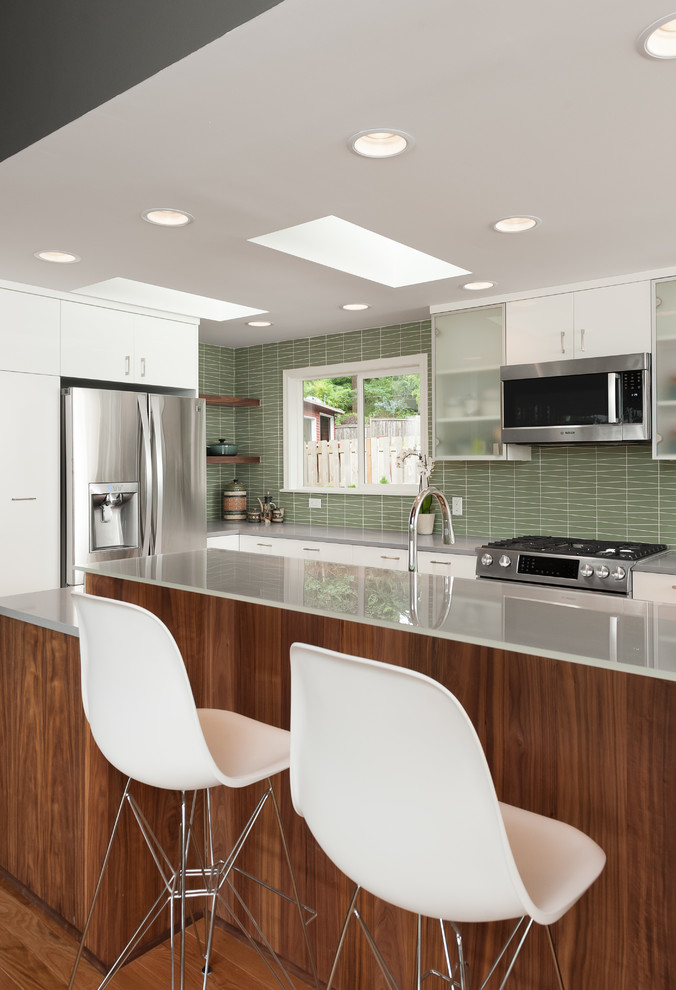 Mid-sized midcentury eat-in kitchen in Portland with a farmhouse sink, flat-panel cabinets, medium wood cabinets, quartz benchtops, green splashback, glass tile splashback, stainless steel appliances, medium hardwood floors, with island, brown floor and grey benchtop.