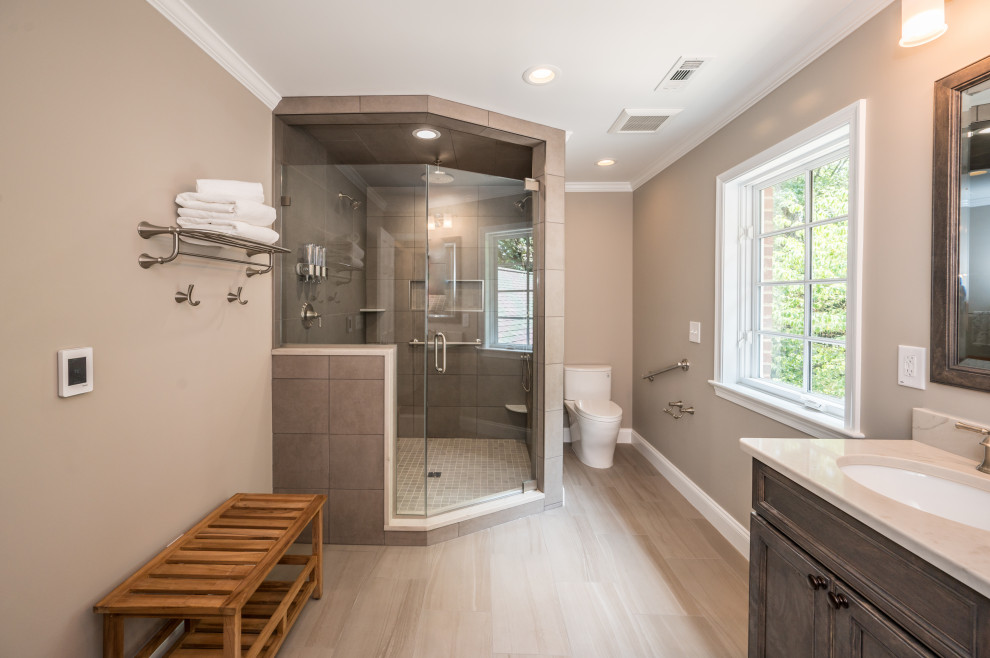 This is an example of a large transitional master bathroom in DC Metro with recessed-panel cabinets, brown cabinets, a corner shower, a two-piece toilet, ceramic floors, an undermount sink, marble benchtops, an open shower, white benchtops, a double vanity and a built-in vanity.