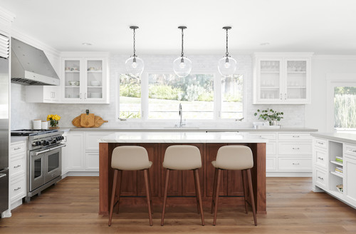 Bright white kitchen with Shaker RTA cabinets, glass-front uppers, wide windows, a walnut island with three stools, and globe pendants on light wood floors.