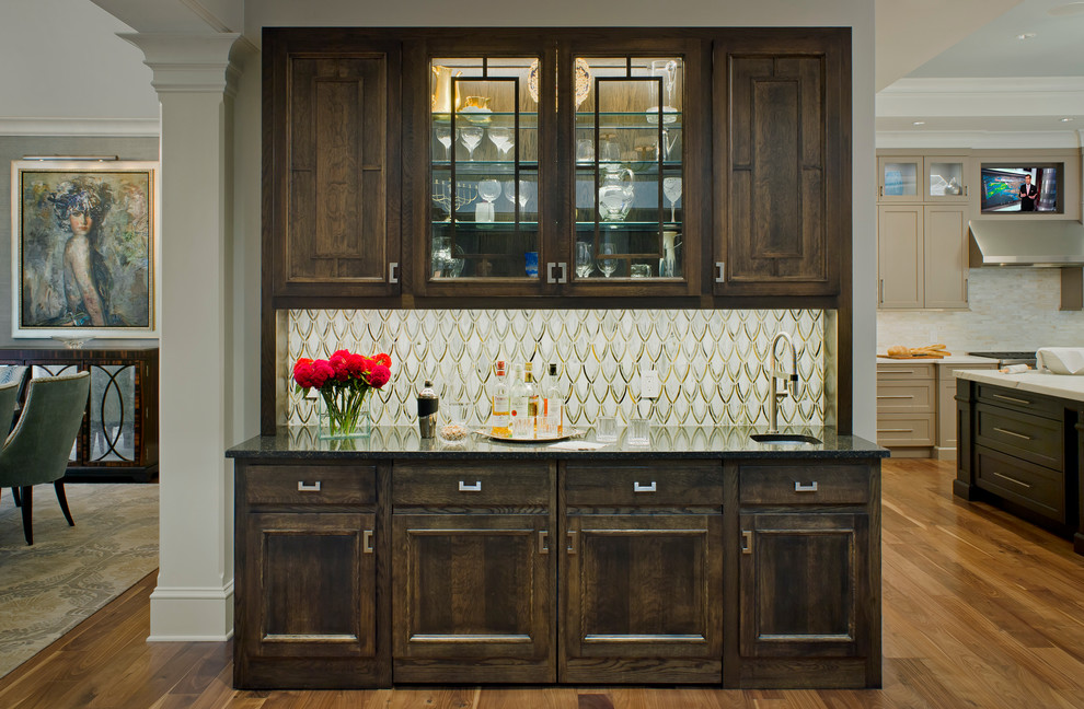 This is an example of a large traditional single-wall wet bar in Baltimore with medium hardwood floors, a drop-in sink, raised-panel cabinets, dark wood cabinets, laminate benchtops, multi-coloured splashback, brown floor and black benchtop.