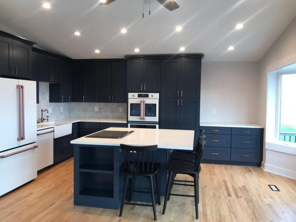 Photo of a mid-sized contemporary l-shaped open plan kitchen in DC Metro with a farmhouse sink, recessed-panel cabinets, blue cabinets, quartzite benchtops, multi-coloured splashback, ceramic splashback, white appliances, light hardwood floors, with island, brown floor and white benchtop.