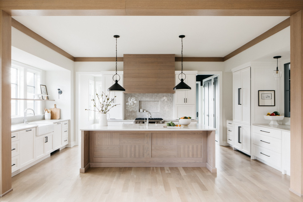 Transitional u-shaped kitchen in Chicago with a farmhouse sink, shaker cabinets, white cabinets, grey splashback, panelled appliances, light hardwood floors, with island, beige floor and white benchtop.