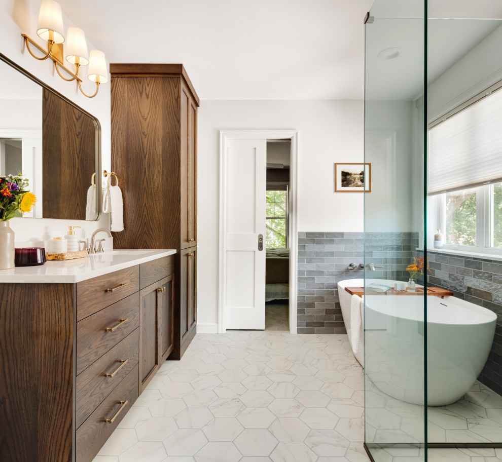 This is an example of a mid-sized transitional master bathroom in Minneapolis with shaker cabinets, medium wood cabinets, a freestanding tub, a curbless shower, gray tile, white walls, porcelain floors, a drop-in sink, engineered quartz benchtops, white floor, a hinged shower door, white benchtops, an enclosed toilet and a built-in vanity.