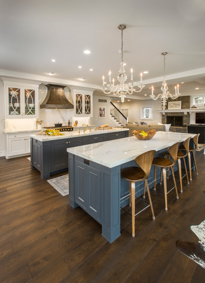 This is an example of a large traditional l-shaped kitchen in Cincinnati with a farmhouse sink, glass-front cabinets, white cabinets, quartz benchtops, white splashback, stone slab splashback, panelled appliances, dark hardwood floors and multiple islands.