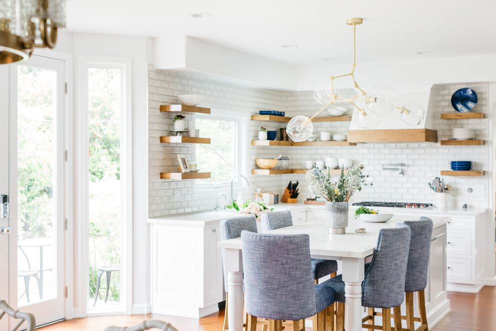 Example of a mid-sized transitional u-shaped medium tone wood floor and brown floor eat-in kitchen design in Los Angeles with a farmhouse sink, recessed-panel cabinets, white cabinets, quartzite countertops, white backsplash, subway tile backsplash, stainless steel appliances, an island and white countertops