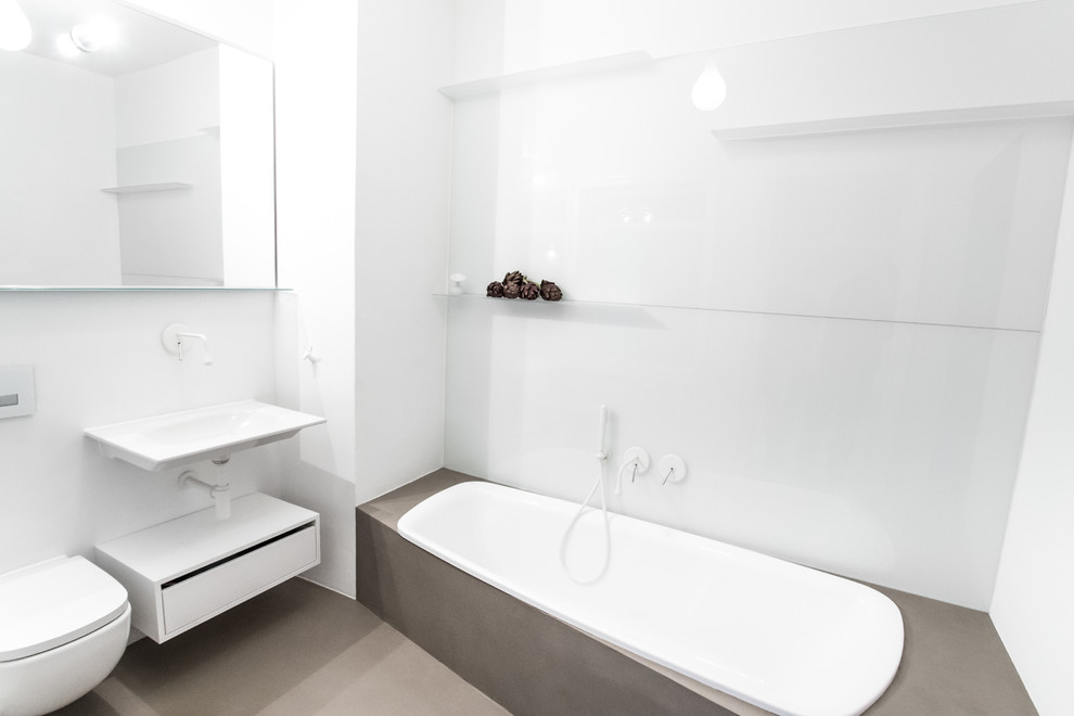 This is an example of a small modern bathroom in Other with a wall-mount sink, flat-panel cabinets, white cabinets, a drop-in tub, a wall-mount toilet, white walls, white tile, glass sheet wall and concrete floors.