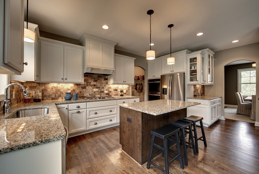 Photo of a large traditional u-shaped kitchen in Minneapolis with an undermount sink, recessed-panel cabinets, white cabinets, granite benchtops, brown splashback, stone tile splashback, stainless steel appliances, dark hardwood floors and with island.