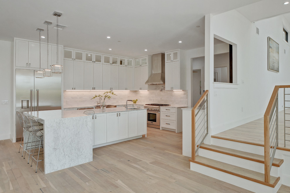 Inspiration for a large transitional l-shaped open plan kitchen in Nashville with a farmhouse sink, shaker cabinets, white cabinets, marble benchtops, white splashback, stainless steel appliances, light hardwood floors and with island.