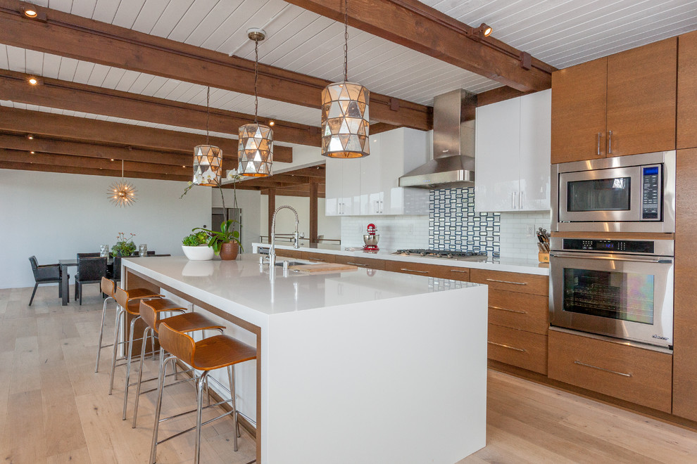 This is an example of a mid-sized midcentury galley open plan kitchen in Orange County with flat-panel cabinets, medium wood cabinets, quartz benchtops, white splashback, glass tile splashback, stainless steel appliances, with island, an undermount sink and light hardwood floors.