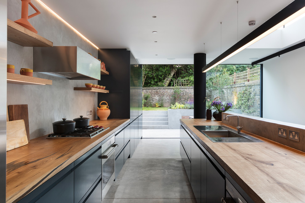 This is an example of a large contemporary galley open plan kitchen in London with flat-panel cabinets, blue cabinets, wood benchtops, grey splashback, with island, a drop-in sink, panelled appliances, concrete floors, grey floor and brown benchtop.