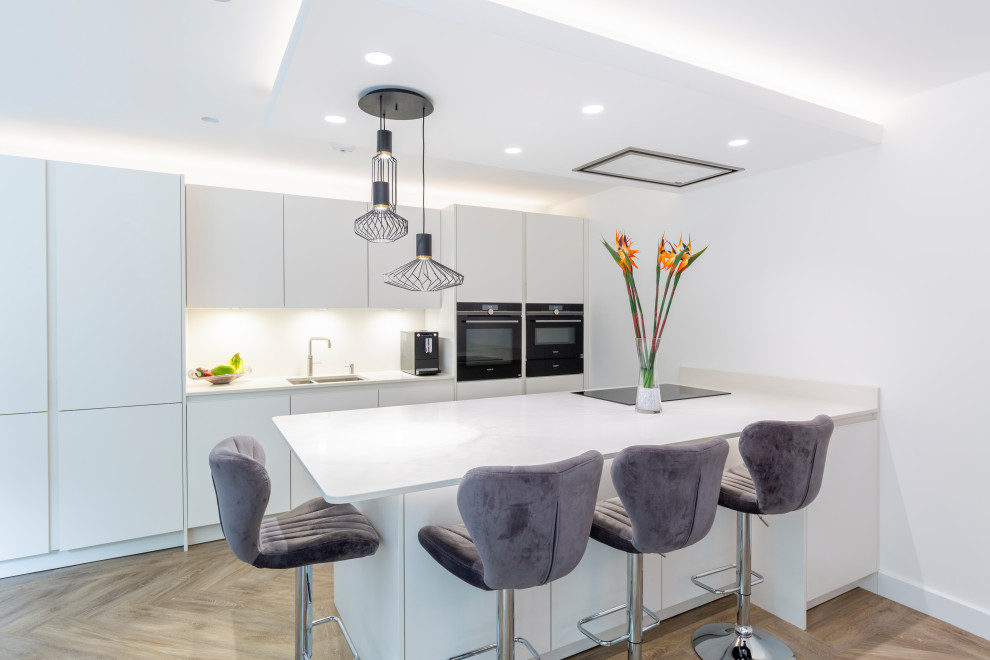 Photo of a medium sized contemporary galley open plan kitchen in Cheshire with a double-bowl sink, flat-panel cabinets, white cabinets, quartz worktops, white splashback, black appliances, an island, brown floors, white worktops, a drop ceiling and feature lighting.