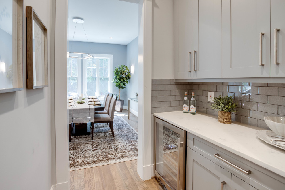This is an example of a mid-sized contemporary single-wall wet bar in DC Metro with shaker cabinets, quartz benchtops, grey splashback, subway tile splashback, medium hardwood floors, brown floor, white benchtop, no sink and grey cabinets.