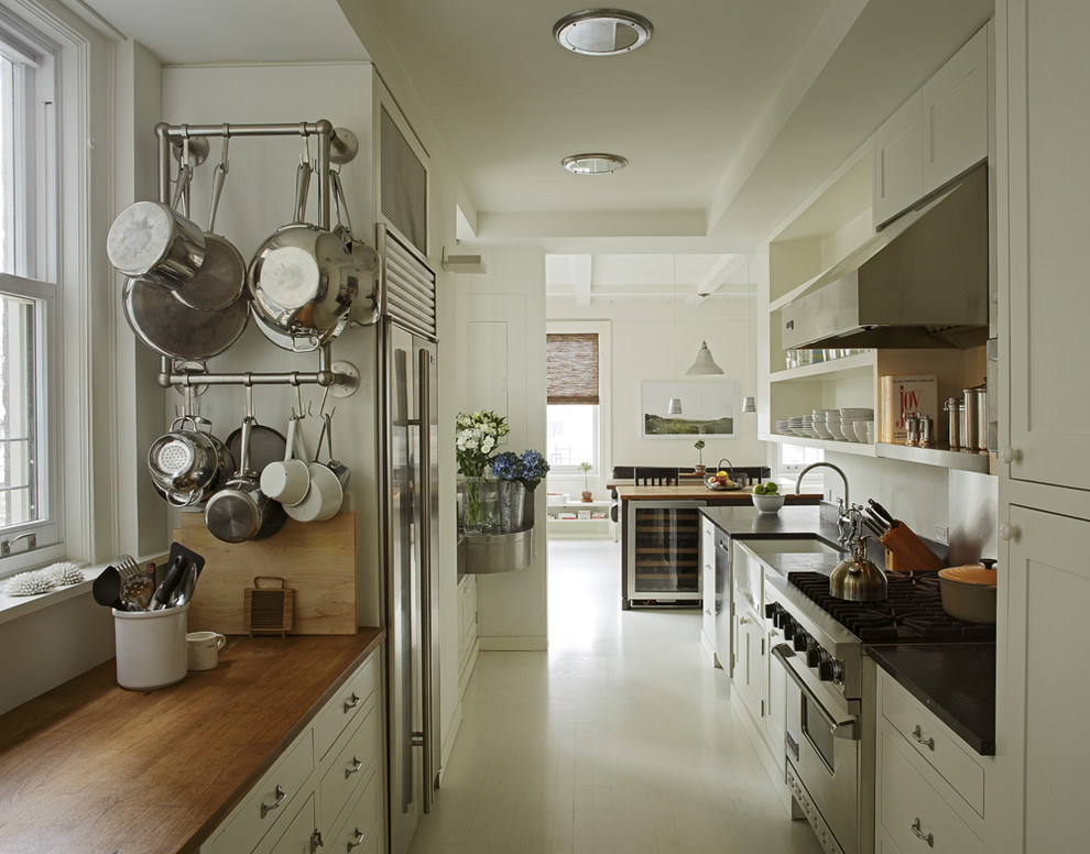 Photo of a modern galley separate kitchen in New York with wood benchtops, a farmhouse sink, open cabinets, white cabinets, stainless steel appliances and white floor.