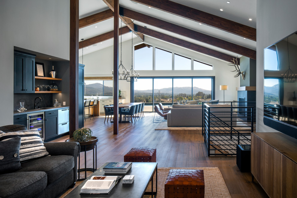 Photo of a large transitional open concept living room in San Francisco with a home bar, grey walls, light hardwood floors, a standard fireplace, a plaster fireplace surround, a wall-mounted tv, brown floor and exposed beam.