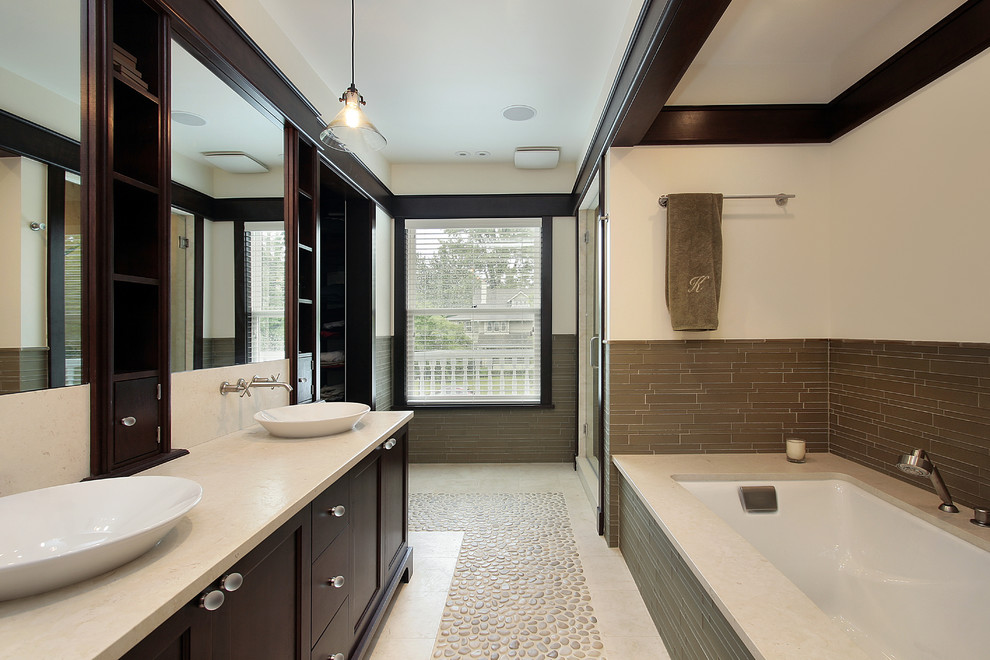 Photo of a large asian master bathroom in Minneapolis with shaker cabinets, brown cabinets, a drop-in tub, an alcove shower, beige walls, pebble tile floors, a vessel sink, limestone benchtops, beige floor and a hinged shower door.