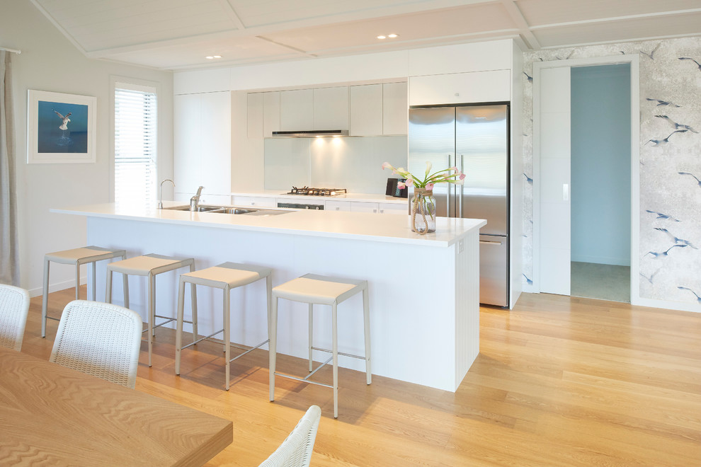 Photo of a mid-sized beach style galley eat-in kitchen in Auckland with a double-bowl sink, white cabinets, quartz benchtops, blue splashback, glass sheet splashback, stainless steel appliances, light hardwood floors and with island.