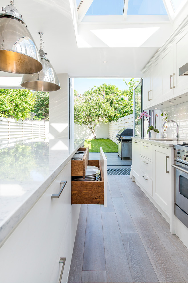 Large contemporary single-wall open plan kitchen in London with a drop-in sink, recessed-panel cabinets, white cabinets, marble benchtops, white splashback, subway tile splashback, stainless steel appliances, light hardwood floors and with island.