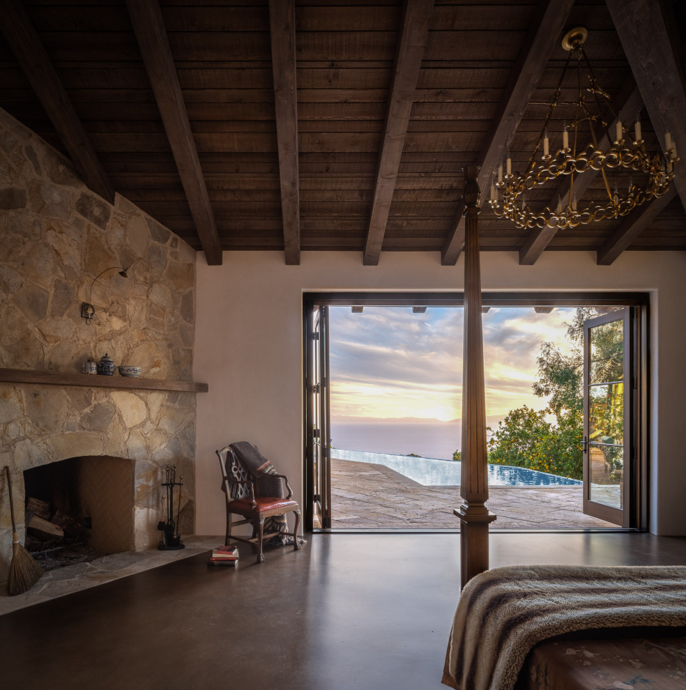 Photo of a large rustic master bedroom in Los Angeles with white walls, concrete flooring, a stone fireplace surround, brown floors, a corner fireplace and exposed beams.