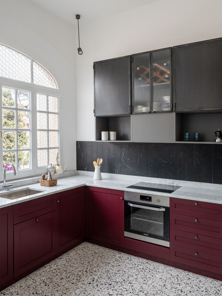 Пример оригинального дизайна: угловая кухня в современном стиле с врезной мойкой, фасадами в стиле шейкер, красными фасадами, черным фартуком, техникой из нержавеющей стали, полом из терраццо, разноцветным полом и белой столешницей