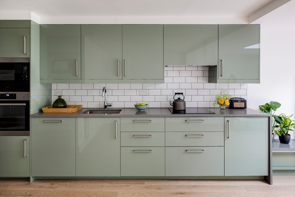 Contemporary single-wall kitchen in London with an undermount sink, flat-panel cabinets, green cabinets, white splashback, subway tile splashback, light hardwood floors, beige floor and grey benchtop.