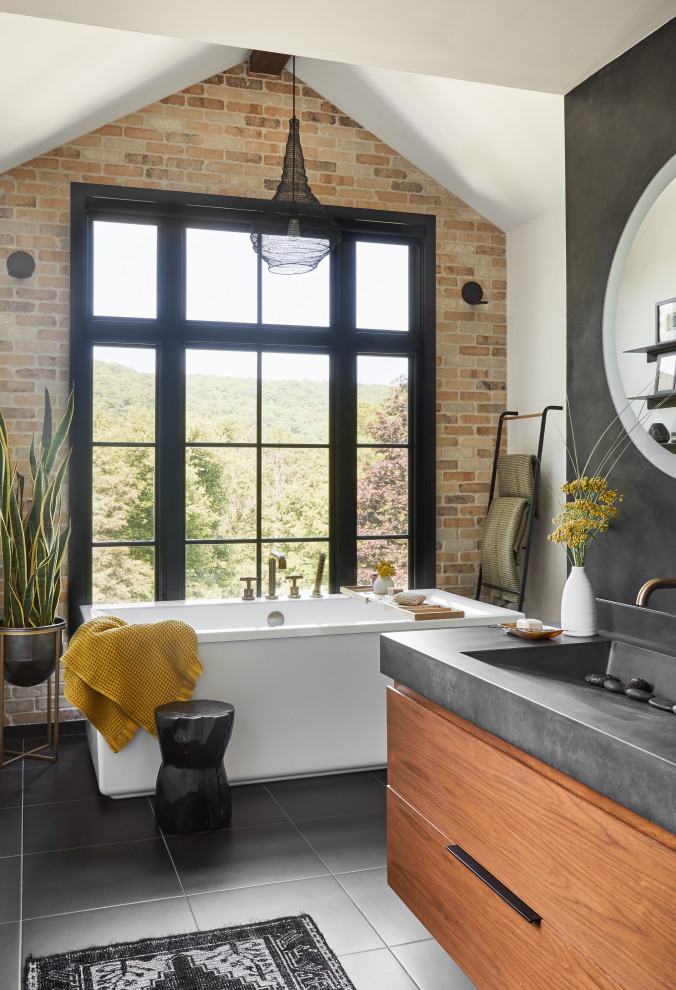 This is an example of a scandinavian bathroom in New York with flat-panel cabinets, medium wood cabinets, a freestanding tub, white walls, an integrated sink, black floor, grey benchtops, a single vanity, a floating vanity, vaulted and brick walls.