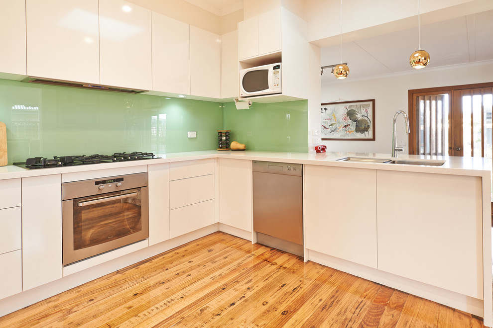 This is an example of a mid-sized contemporary u-shaped kitchen in Melbourne with a double-bowl sink, flat-panel cabinets, white cabinets, quartz benchtops, green splashback, glass sheet splashback, stainless steel appliances and medium hardwood floors.