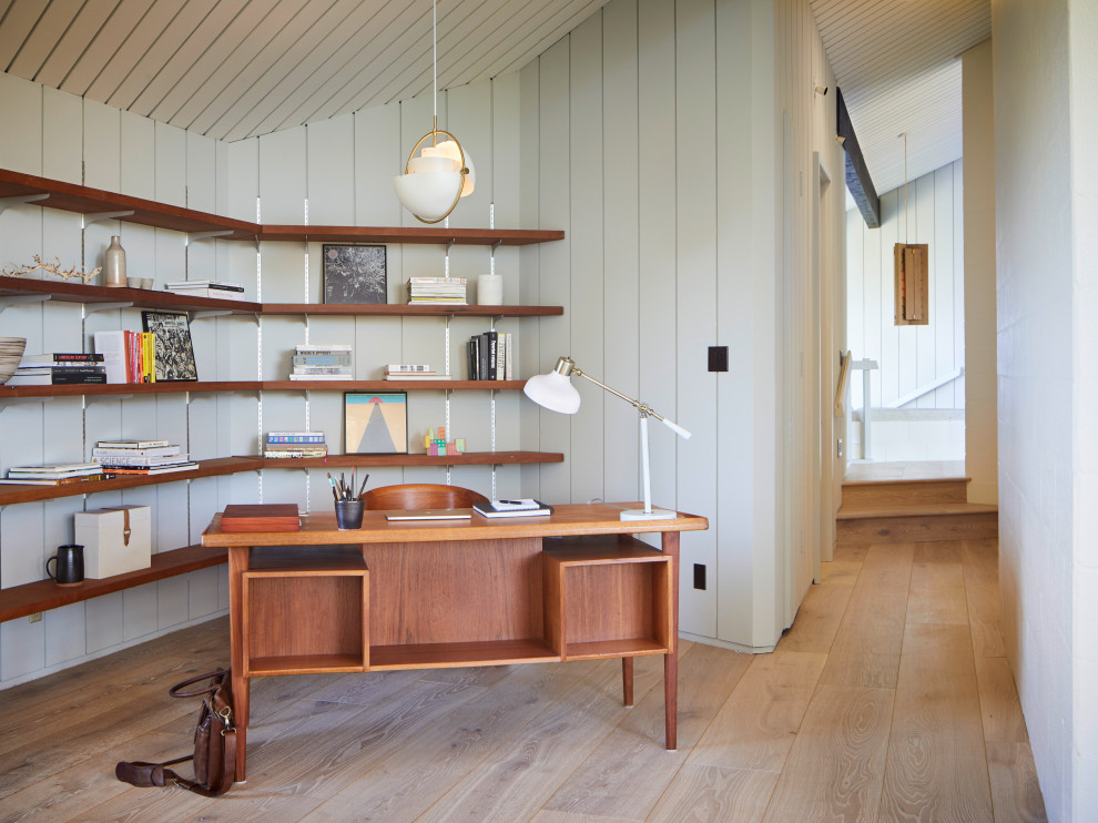 Photo of a large midcentury home office in San Francisco with a library, grey walls, light hardwood floors, a freestanding desk, exposed beam and panelled walls.