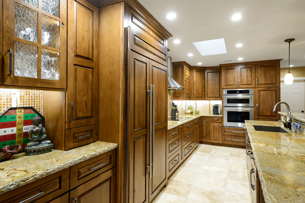 Example of a mid-sized mountain style l-shaped travertine floor open concept kitchen design in Phoenix with a drop-in sink, raised-panel cabinets, medium tone wood cabinets, granite countertops, green backsplash, mosaic tile backsplash, paneled appliances and an island