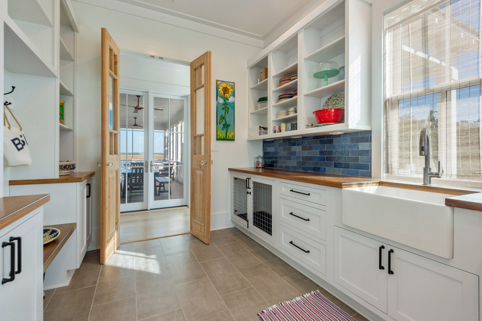 Mid-sized transitional l-shaped utility room in New Orleans with a farmhouse sink, shaker cabinets, white cabinets, wood benchtops, blue splashback, glass tile splashback, white walls, ceramic floors, beige floor and brown benchtop.