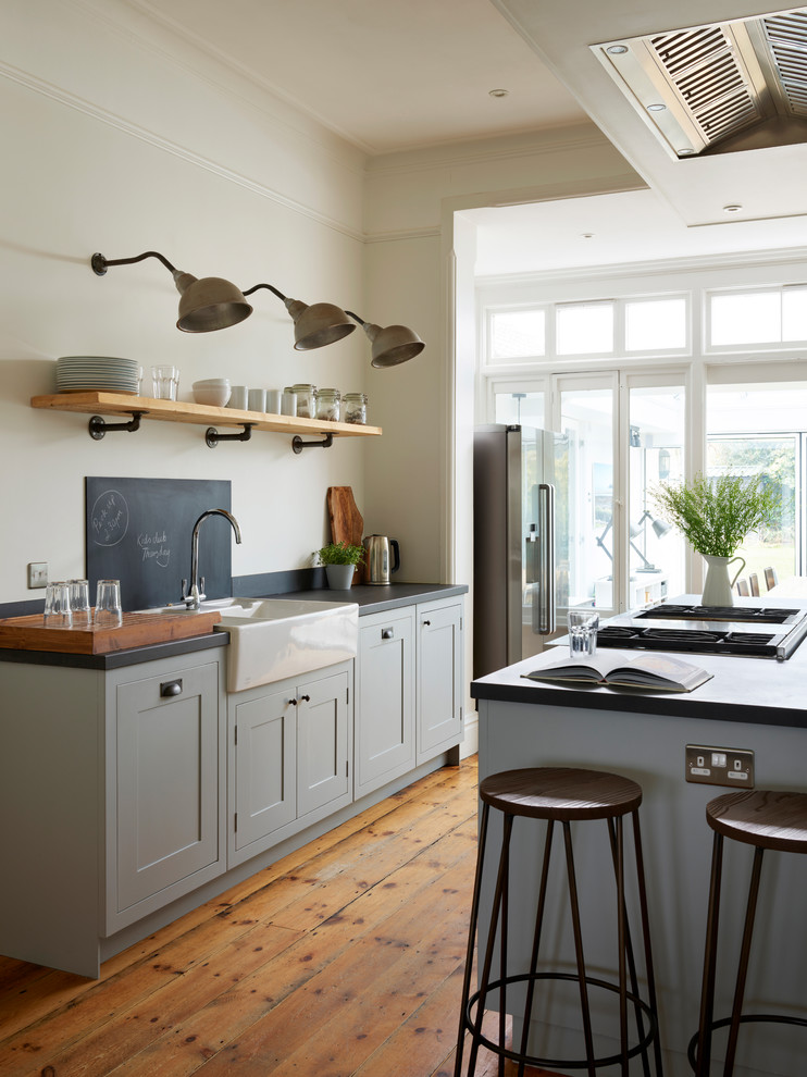 Photo of a large classic open plan kitchen in Essex with a belfast sink, shaker cabinets, grey cabinets, black splashback, medium hardwood flooring, an island, brown floors and black worktops.