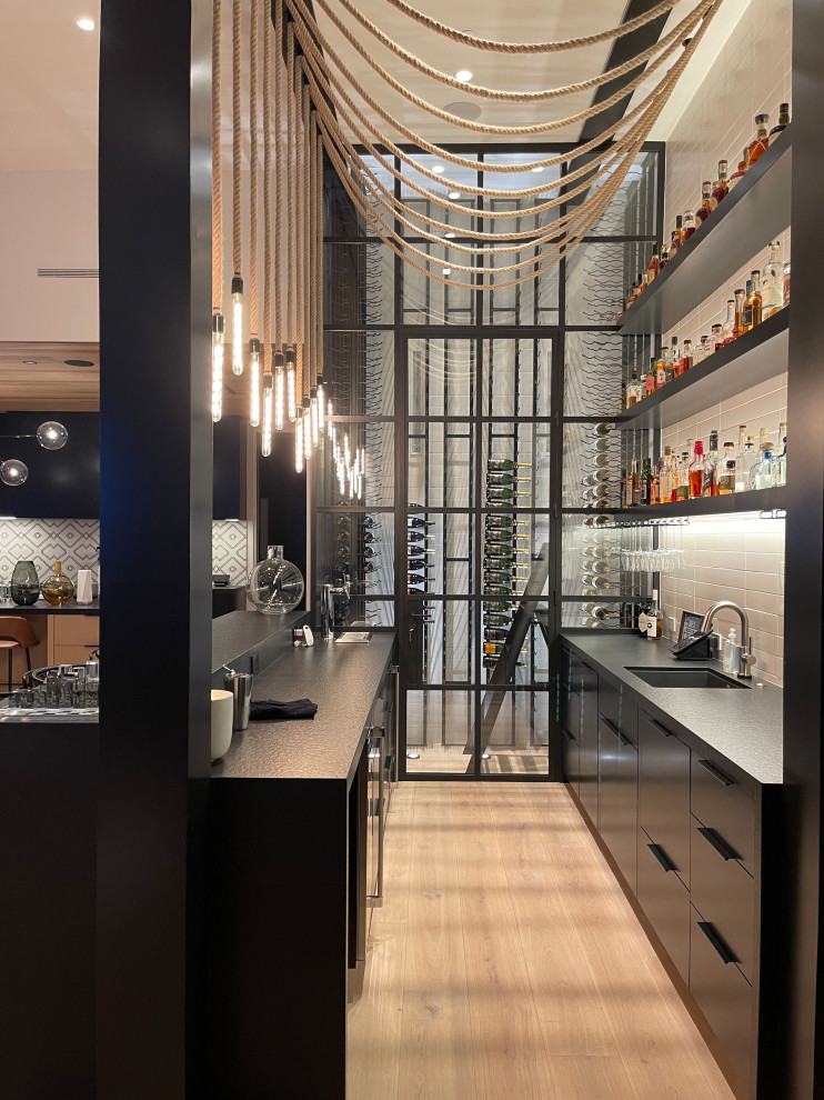 Small contemporary single-wall home bar in Orange County with an integrated sink, flat-panel cabinets, dark wood cabinets, white splashback, subway tile splashback, light hardwood floors, beige floor and black benchtop.