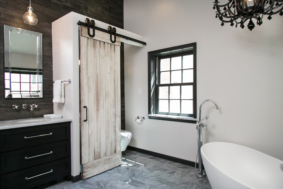 Design ideas for a mid-sized eclectic master bathroom in Philadelphia with an undermount sink, recessed-panel cabinets, black cabinets, marble benchtops, a freestanding tub, a curbless shower, a wall-mount toilet, gray tile, stone tile, white walls and marble floors.