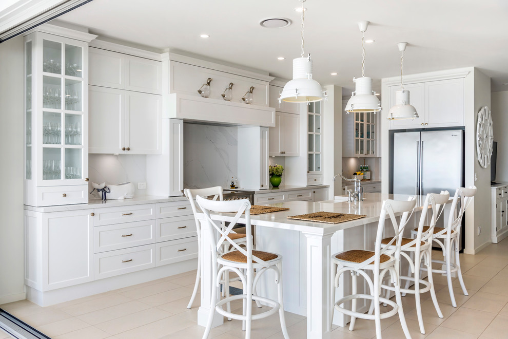 Beach style l-shaped kitchen in Gold Coast - Tweed with a farmhouse sink, shaker cabinets, white cabinets, white splashback, stainless steel appliances, with island, beige floor and white benchtop.