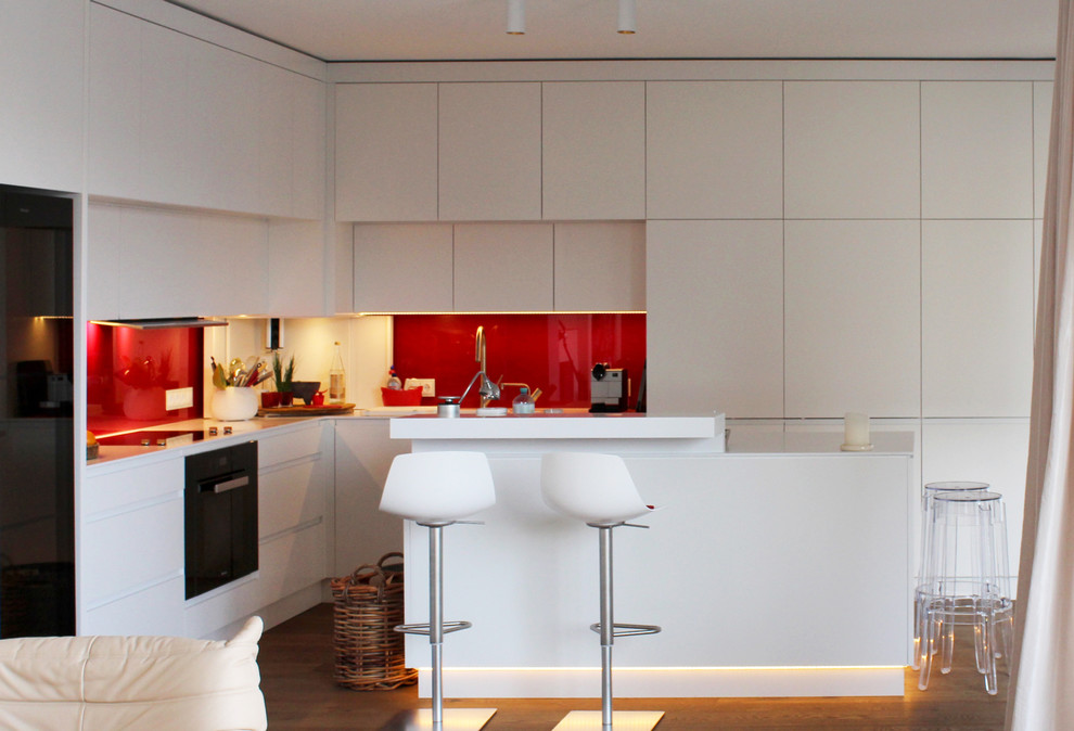 This is an example of a mid-sized contemporary l-shaped open plan kitchen in Berlin with a double-bowl sink, flat-panel cabinets, white cabinets, solid surface benchtops, red splashback, glass sheet splashback, black appliances, painted wood floors and with island.