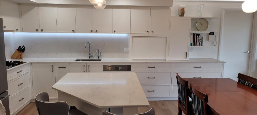 Photo of a mid-sized contemporary l-shaped eat-in kitchen in Sydney with a single-bowl sink, shaker cabinets, white cabinets, quartz benchtops, white splashback, engineered quartz splashback, stainless steel appliances, laminate floors, with island, brown floor, white benchtop and recessed.
